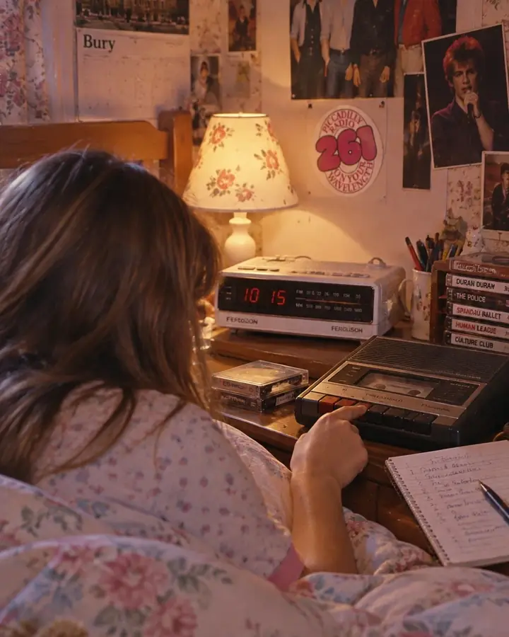Recreated photo: a teenage girl seen from behind under a floral duvet in an early-1980s bedroom in Bury, a white clock radio glowing on the bedside table, her finger on a cassette recorder's record button, pop posters and a Piccadilly Radio 261 sticker on the wall.
