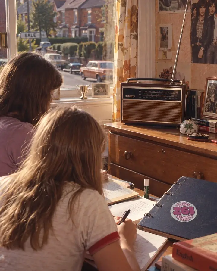 Recreated photo: two teenage sisters seen from behind doing homework in a 1970s bedroom in Sale, a Roberts radio on the dresser and a Piccadilly Radio 261 sticker on a school binder, terraced houses through the window.